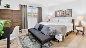 A bedroom with a bed, bedside tables, a desk, a chair, a sofa, a window with curtains, and artwork on the wall.