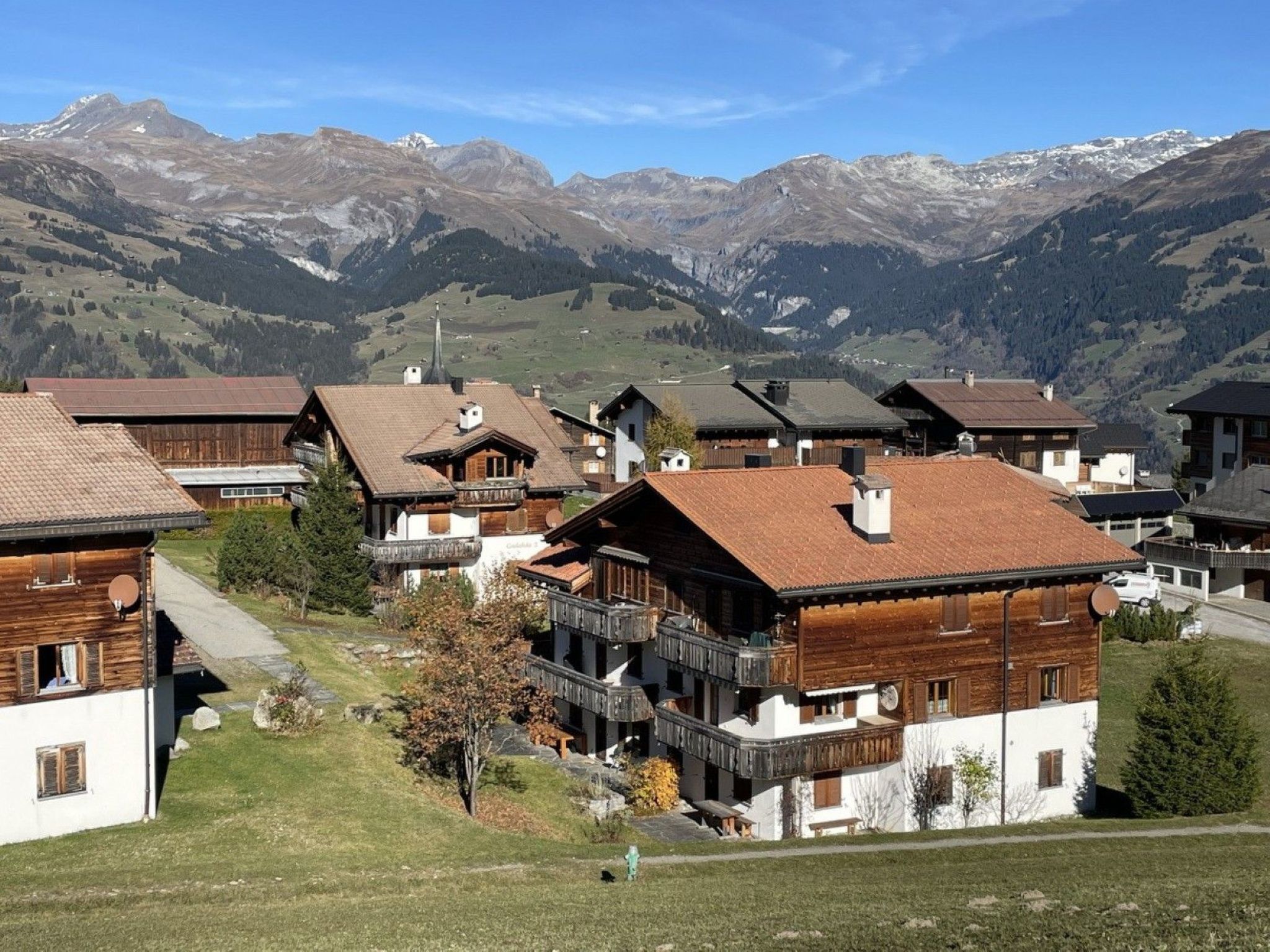A mountain village with traditional wooden houses, a clear sky, and snow-capped mountains in the background.