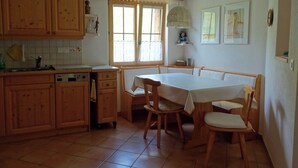 A kitchen with wooden cabinets, a dining table with chairs, a window with curtains, and a hanging light fixture. - Gadahüs Elvers, Obersaxen-Misanenga, (Obersaxen), 34091B by Interhome (Obersaxen Affeier)