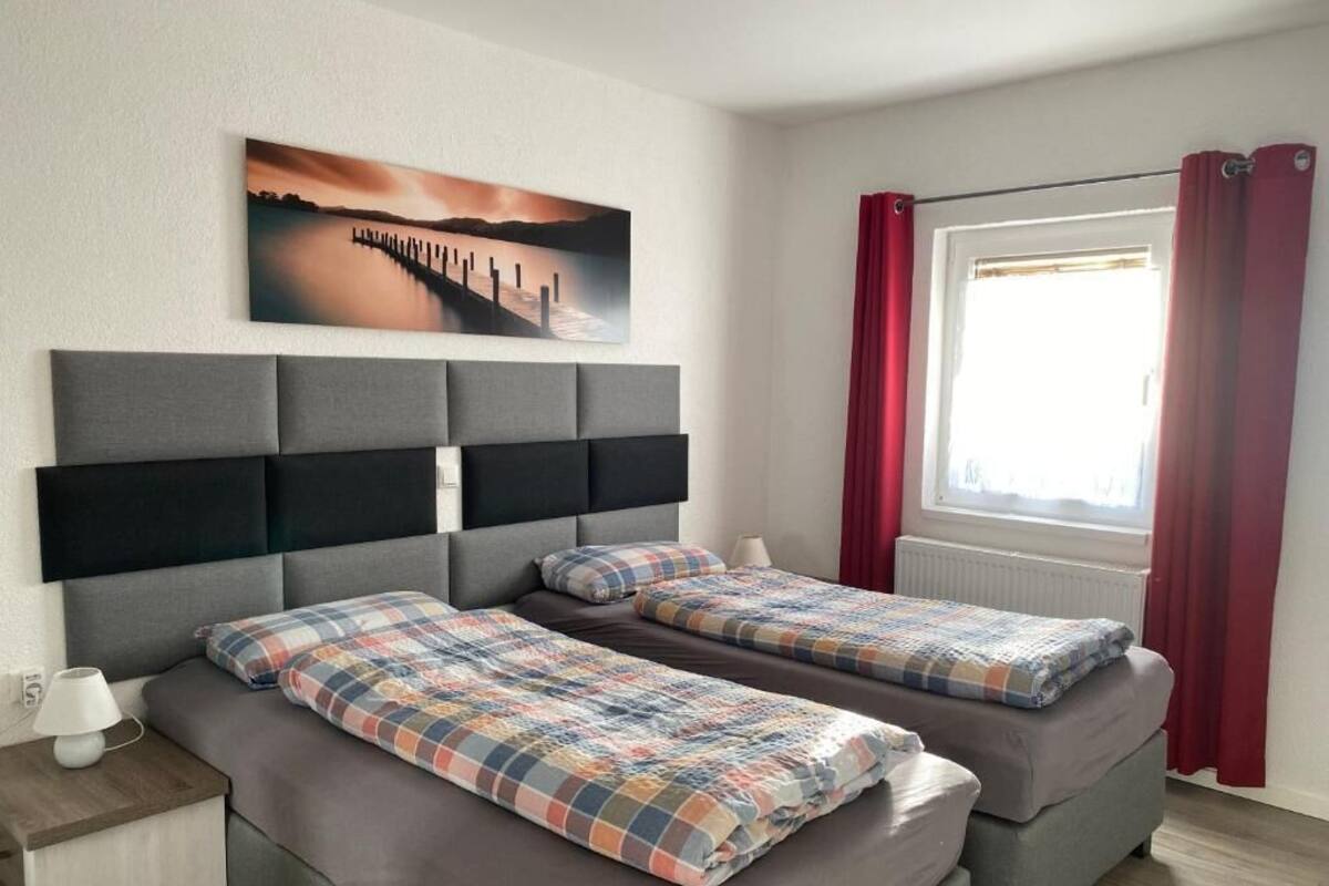 A room with two beds, a headboard, a window with red curtains, and a painting on the wall.