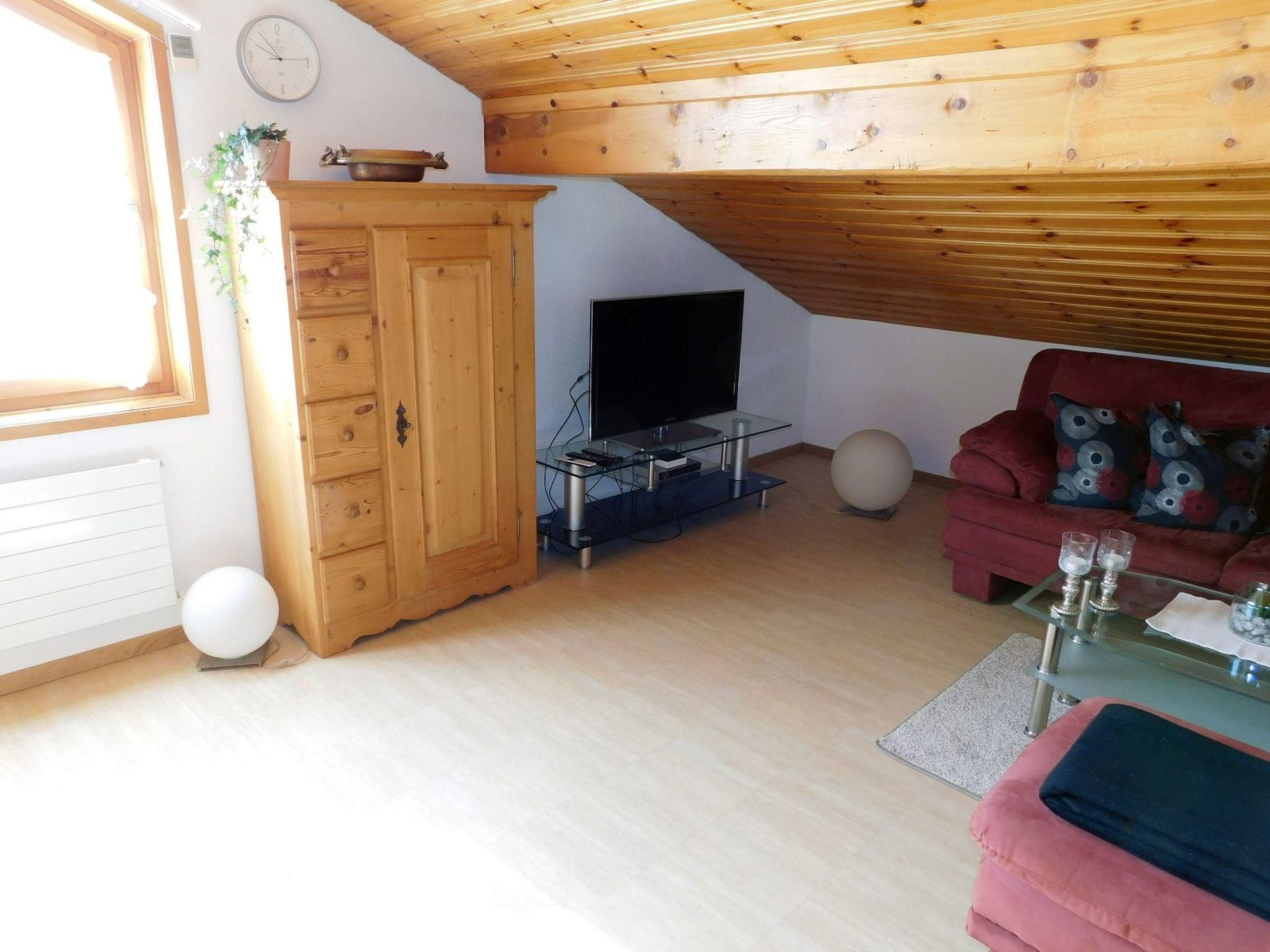 Una sala de estar con techo de madera, un televisor sobre un pedestal de vidrio, un sofá rojo y un mueble de madera.