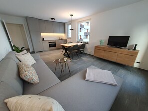 Ein modernes Wohnzimmer mit einer Couch, einem Couchtisch, einem Essbereich mit Stühlen, einer Küche mit Schränken und einem Fernseher auf einer Holzkommode.