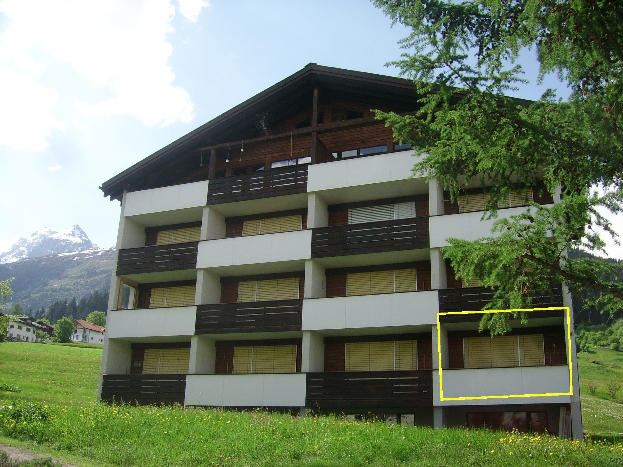 A multi-story building with balconies, set in a green landscape with mountains in the background.