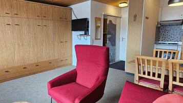 Ein Zimmer mit einem rot gepolsterten Sessel, einem Holztisch, einer Küchenzeile mit Herd und einem Holzschrank.
