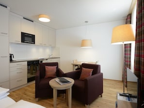 Un espacio habitable compacto con kitchenette, dos sillones, una mesita redonda y comedor.