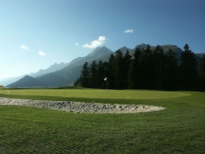 Un campo de golf con un búnker de arena y un mástil de bandera, rodeado de árboles y montañas.