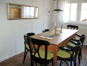 Un comedor con mesa de madera, sillas negras con cojines verdes, un espejo y una lámpara colgante.