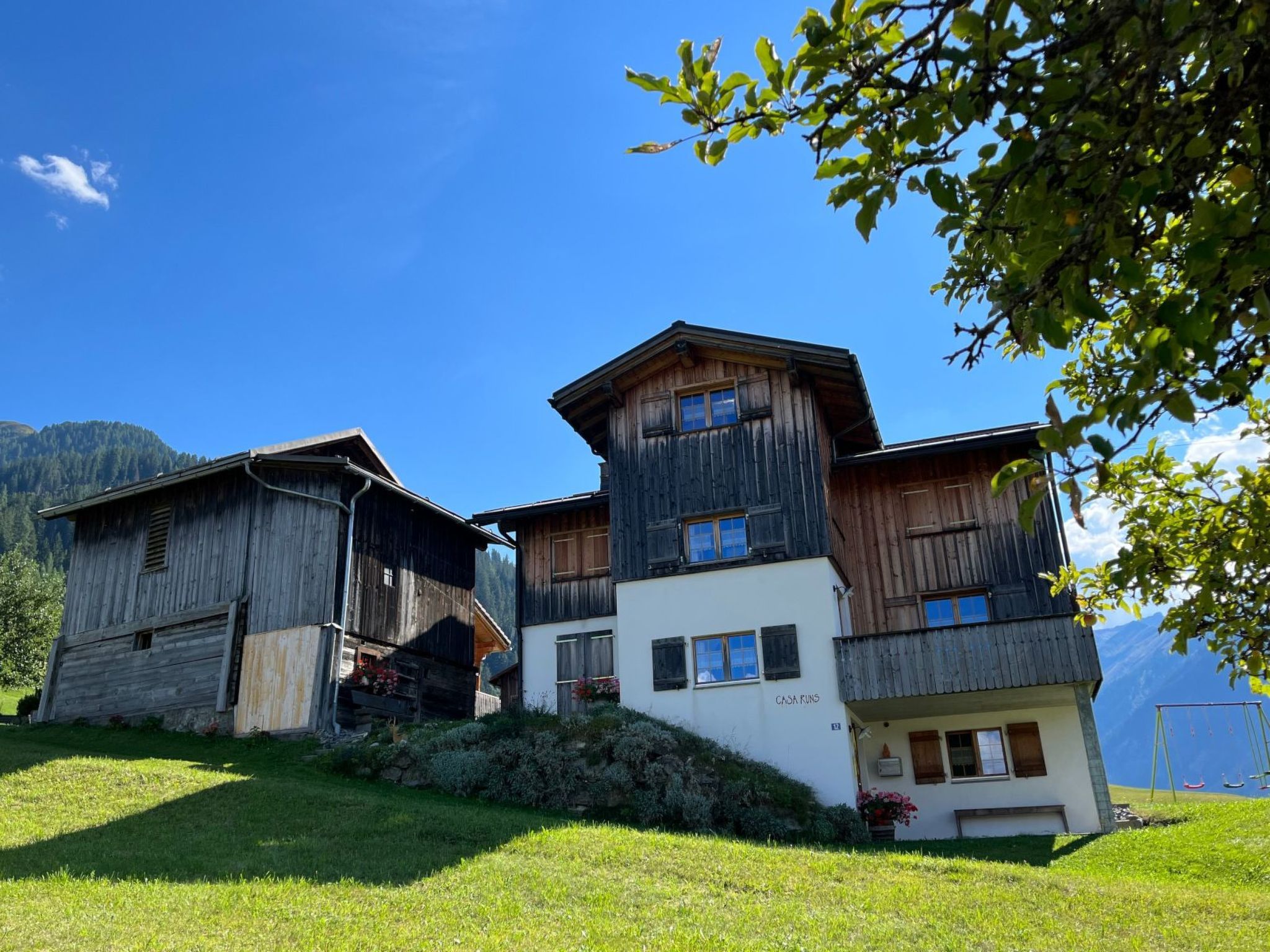 A two-story wooden chalet with a balcony, surrounded by greenery and a clear sky.