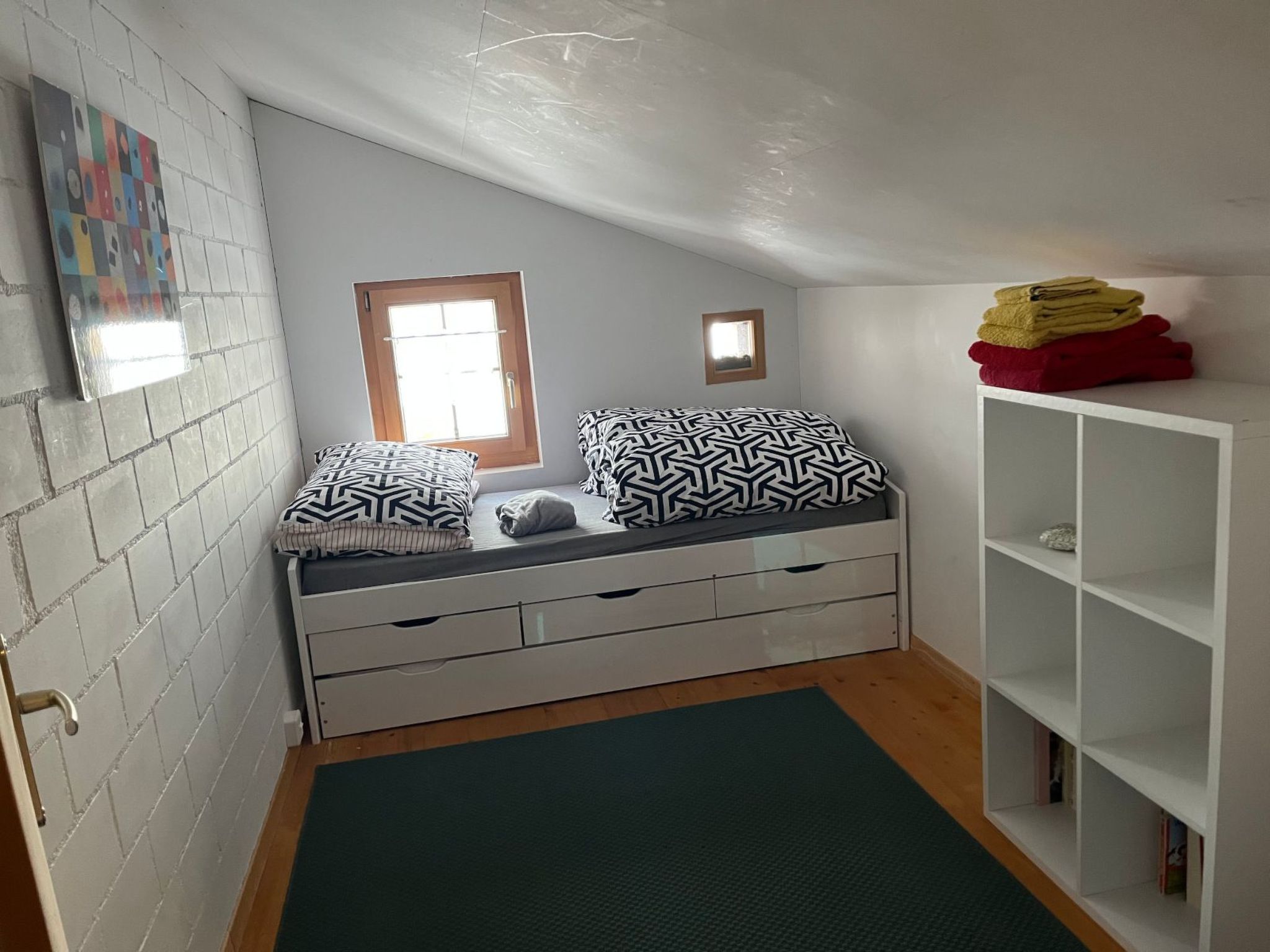 Quarto pequeno com uma cama de solteiro, janela, guarda-roupa branco e tapete verde no chão.