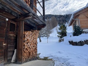 Eine Holzhütte mit verschneiter Fassade, ein Holzstoß und eine verschneite Landschaft mit Bäumen und einem Berg im Hintergrund.
