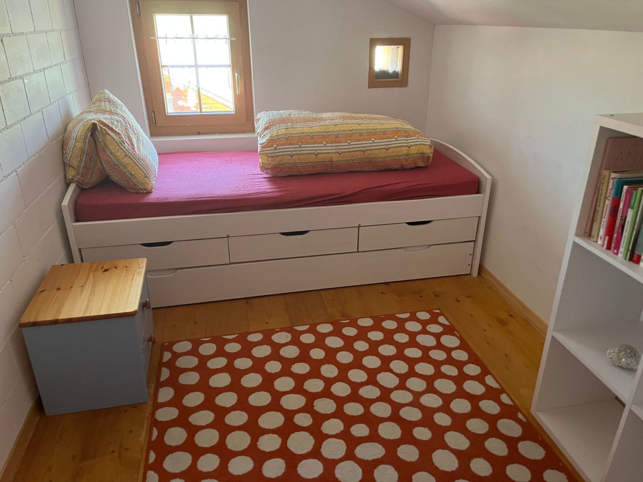 Une petite chambre à coucher avec un lit, une table de chevet en bois, un tapis à pois et une bibliothèque.