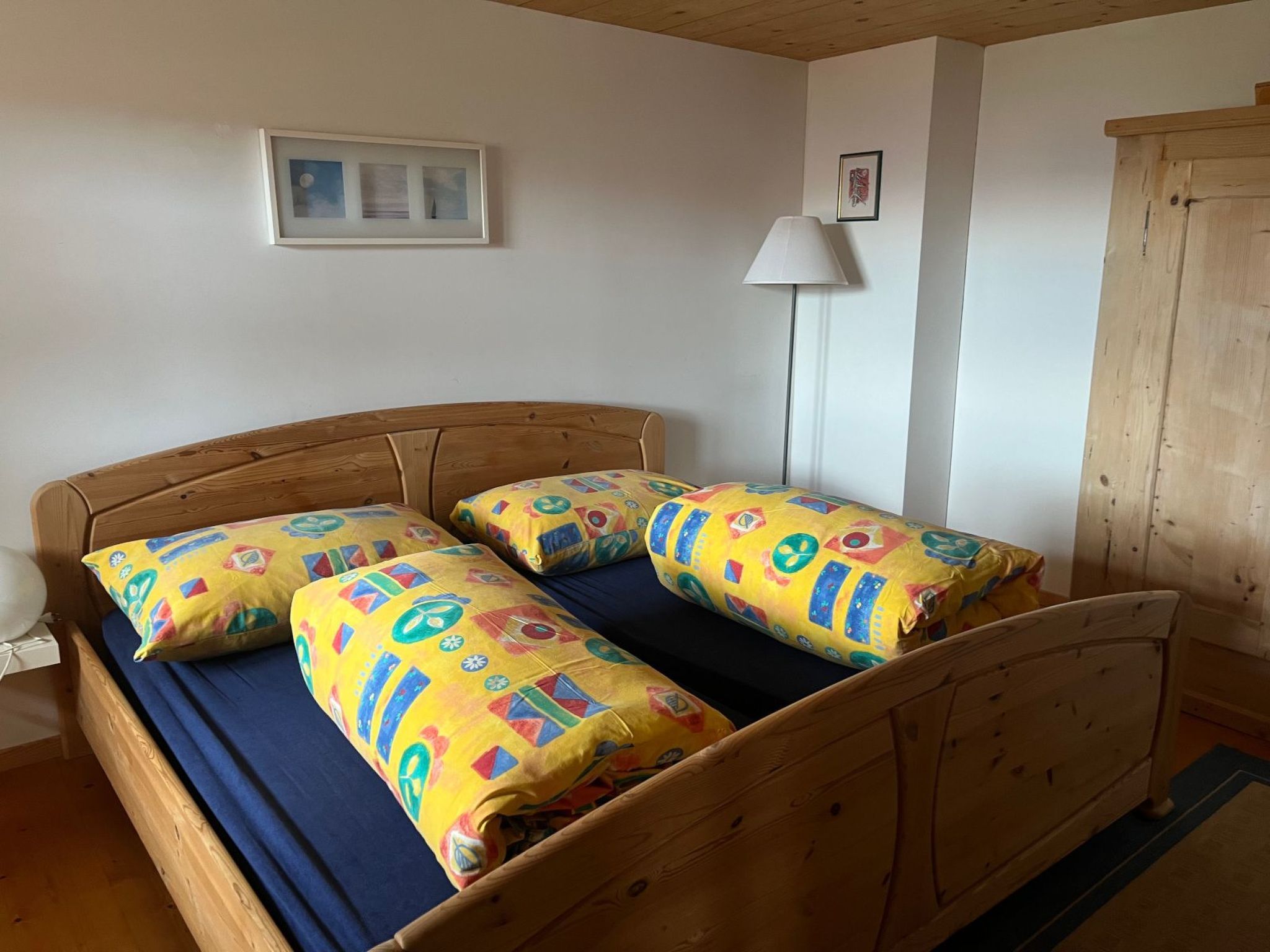 Una cama de madera con ropa de cama azul y almohadas coloridas, una pared blanca con cuadros enmarcados y un armario de madera.