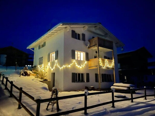 A two-story house with a balcony, surrounded by snow, and decorated with string lights.