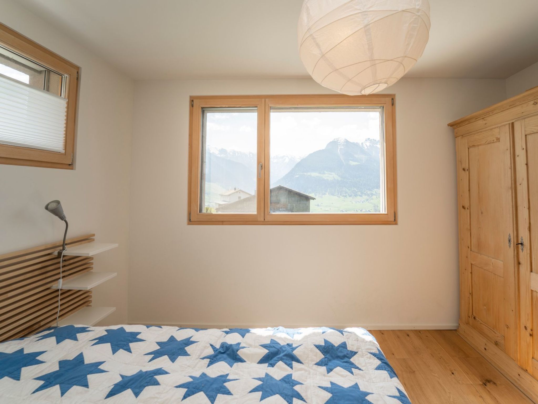 Un dormitorio con una cama, una ventana con vistas a montañas, un armario de madera y una lámpara de noche.
