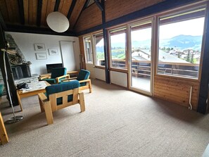 Une pièce avec un plafond en bois, une table entourée de chaises, un téléviseur et de grandes fenêtres donnant sur une ville et des montagnes.