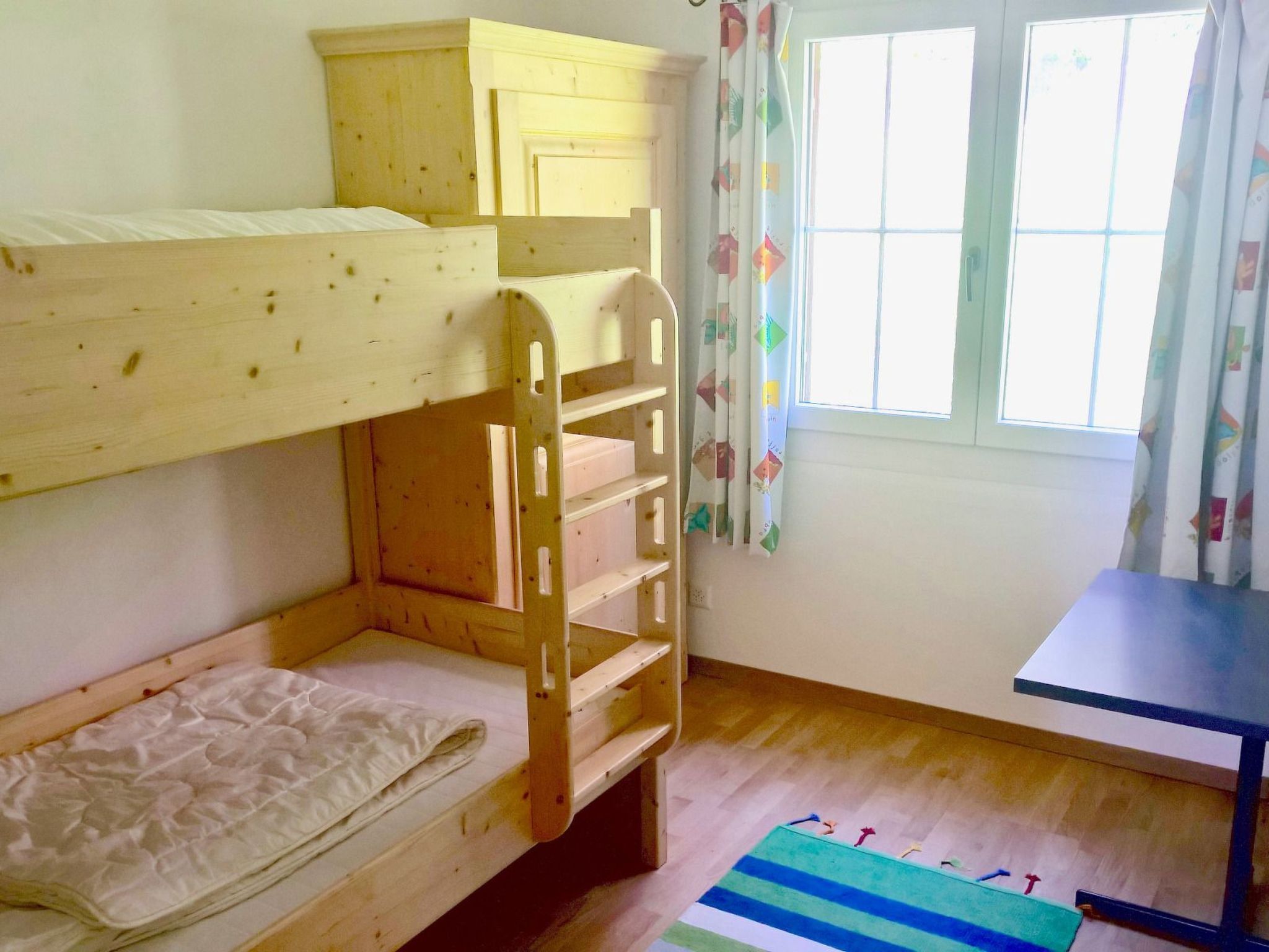 Habitación con literas, ventana con cortinas, suelo de madera y una mesita.