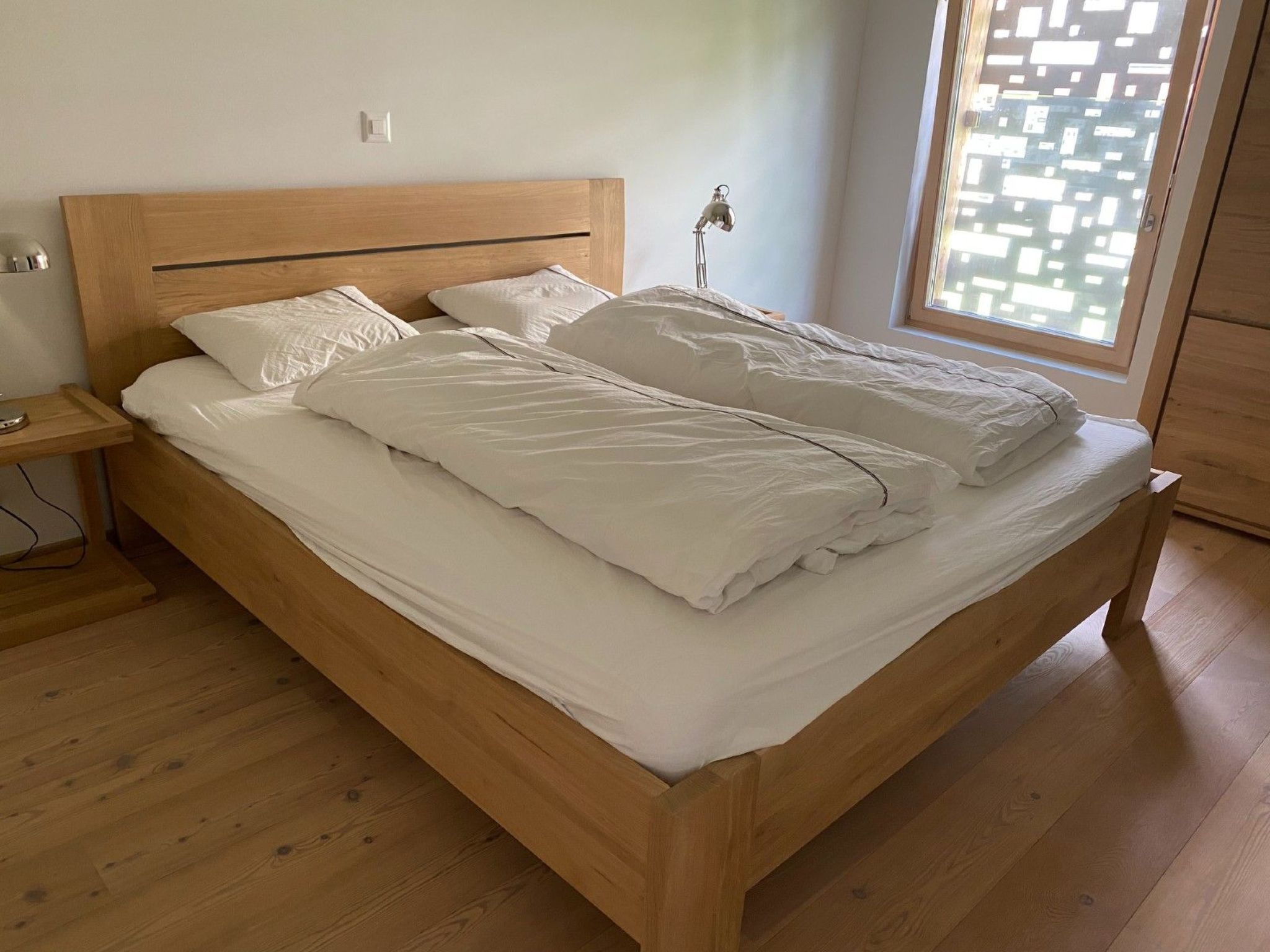 A wooden bed with white bedding in a room with wooden flooring and a window.