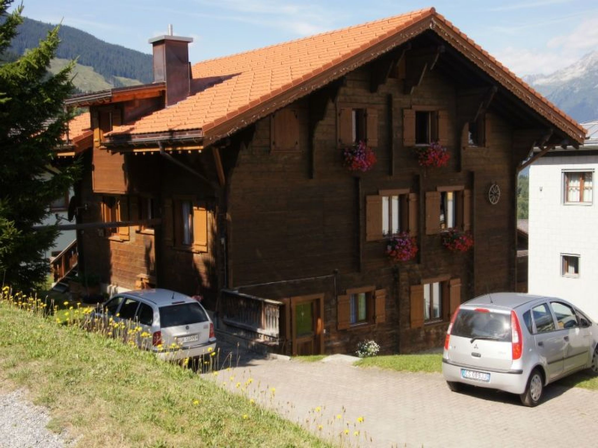A wooden chalet with a sloped roof, multiple windows, and flower boxes. Two cars are parked in front.