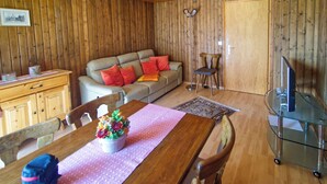 A wooden-paneled room with a sofa, dining table, chairs, a small cabinet, a floor lamp, and a television on a glass stand. - Genoveva Sax, Obersaxen-Meierhof, (Obersaxen), 34065B by Interhome (Obersaxen Affeier)
