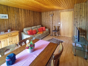 A wooden-paneled room with a sofa, dining table, chairs, a small cabinet, a floor lamp, and a television on a glass stand. - Genoveva Sax, Obersaxen-Meierhof, (Obersaxen), 34065B by Interhome (Obersaxen Affeier)