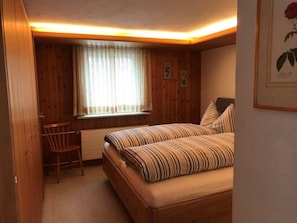 Ein Schlafzimmer mit Holzboden, einem Bett mit gestreifter Bettwäsche, einem Stuhl, einem Fenster mit Vorhängen und einem gerahmten Bild an der Wand.