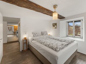 A bedroom with a wooden ceiling beam, a bed with floral bedding, a bedside table with a lamp, and a window with a view of mountains. - Pacifica und Romantica in Cumpadials, (Cumpadials), 76002B by Interhome (Cumpadials)