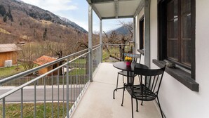 Un balcón con mesa y sillas, vista a un paisaje montañoso y una ventana con vistas a las montañas.
