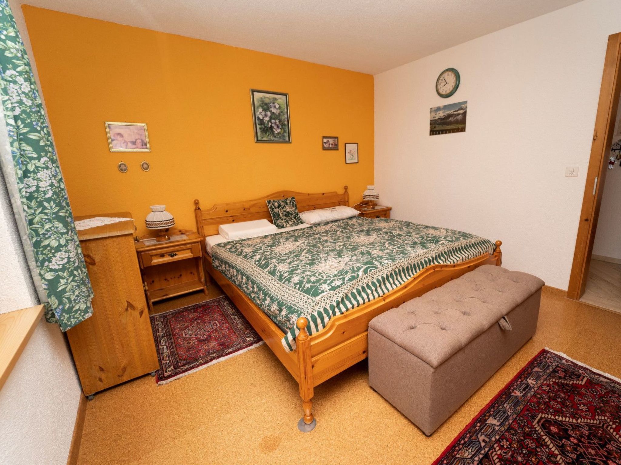 A bedroom with a wooden bed, a bench, a nightstand, a lamp, and a wall clock.
