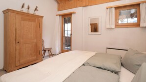 Un dormitorio con un armario de madera, una cama con ropa de cama gris, una ventana pequeña con cortinas y un espejo en la pared.