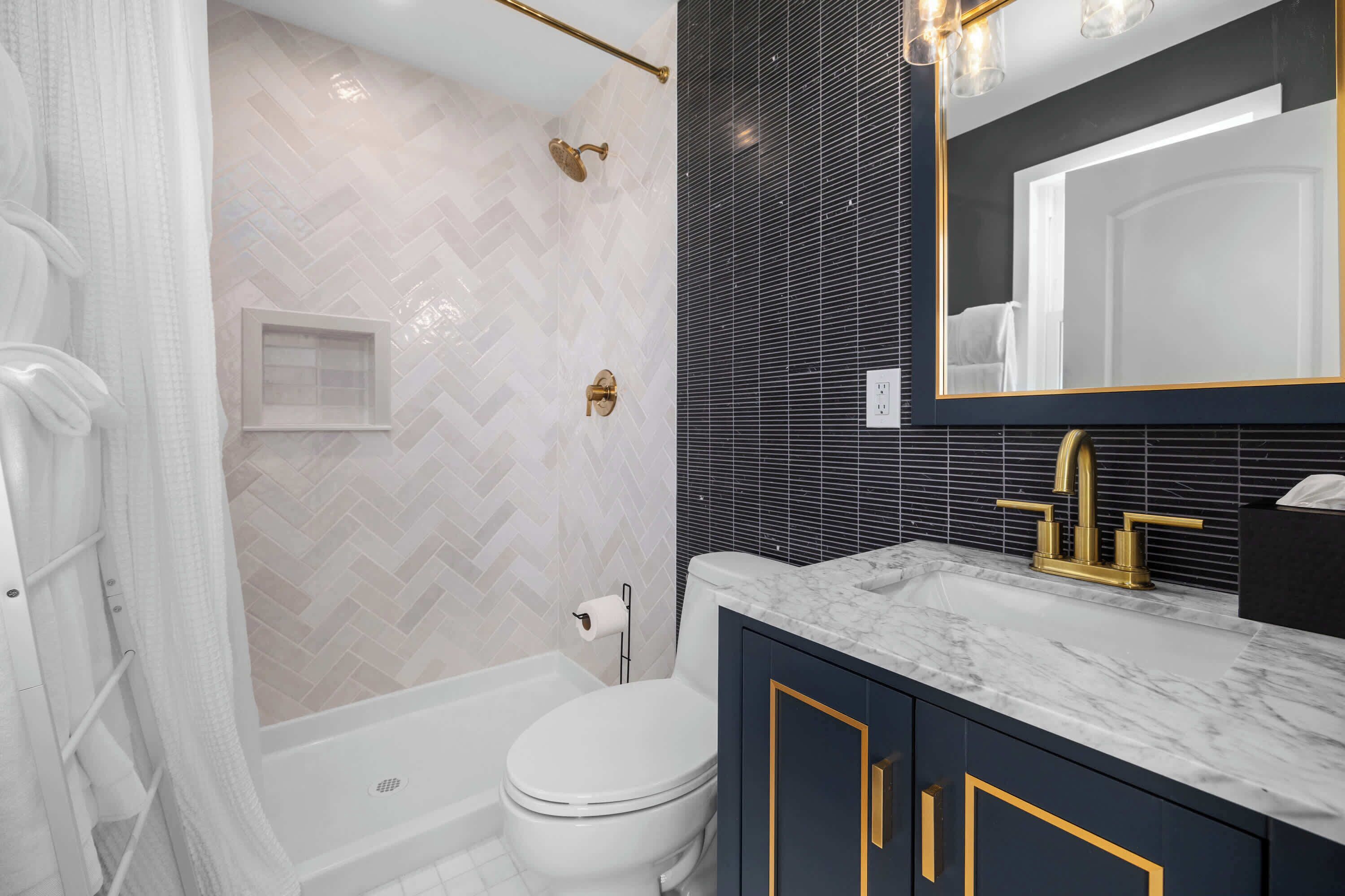 Ein Badezimmer mit einer weißen Toilette, einer Dusche mit goldfarbener Duschbrause und einem dunklen Waschtisch mit goldfarbenem Wasserhahn.
