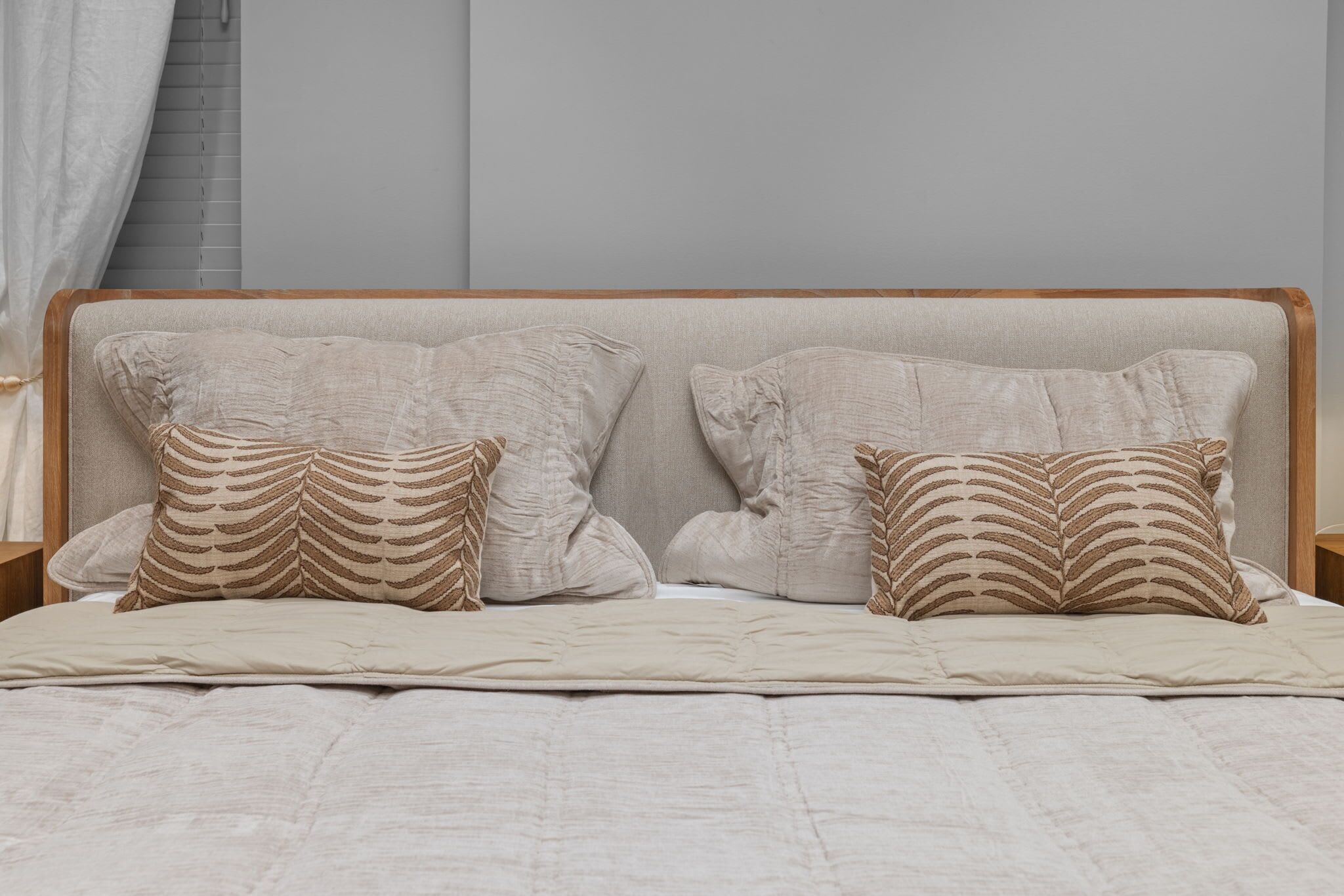 Una cama con cabecera de madera, dos almohadas estampadas y una colcha de color neutro.