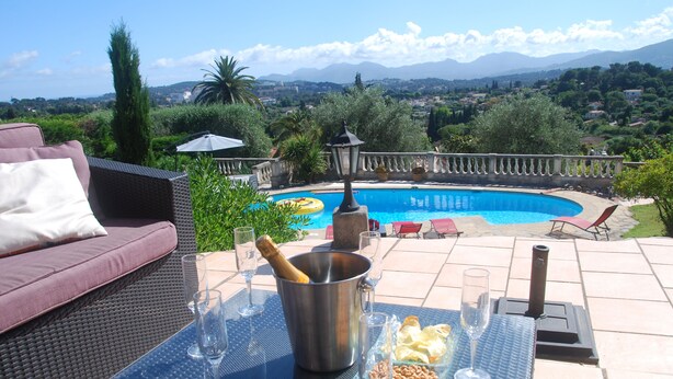 Un espacio junto a la piscina con un sofá de mimbre, una mesa con vasos y aperitivos, y vistas a una piscina y montañas.
