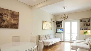 Ein modernes Wohnzimmer mit Glastisch, weißen Stühlen, einem weißen Sofa, einem Fernseher, einem Bücherregal und Kunstwerken an den Wänden.
