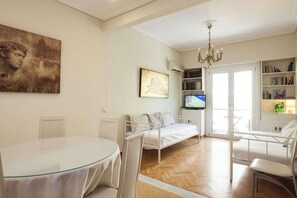 A modern living room with a glass table, white chairs, a white sofa, a TV, a bookshelf, and artwork on the walls. - Lovely 1 Bd Apt with Balcony 10 min from Acropolis (Athina)