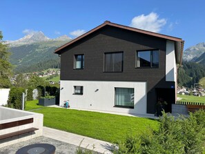 Une maison moderne à deux étages, dotée d’une façade sombre, de grandes fenêtres et offrant une vue sur les montagnes en arrière-plan.