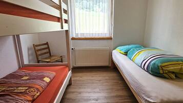 Ein kleiner, schmaler Raum mit zwei Etagenbetten, einem Holzstuhl, einer Heizung und einem Fenster mit Vorhang.
