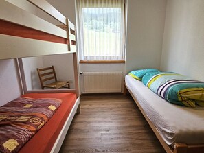 A small, narrow room with two bunk beds, a wooden chair, a radiator, and a window with a curtain.