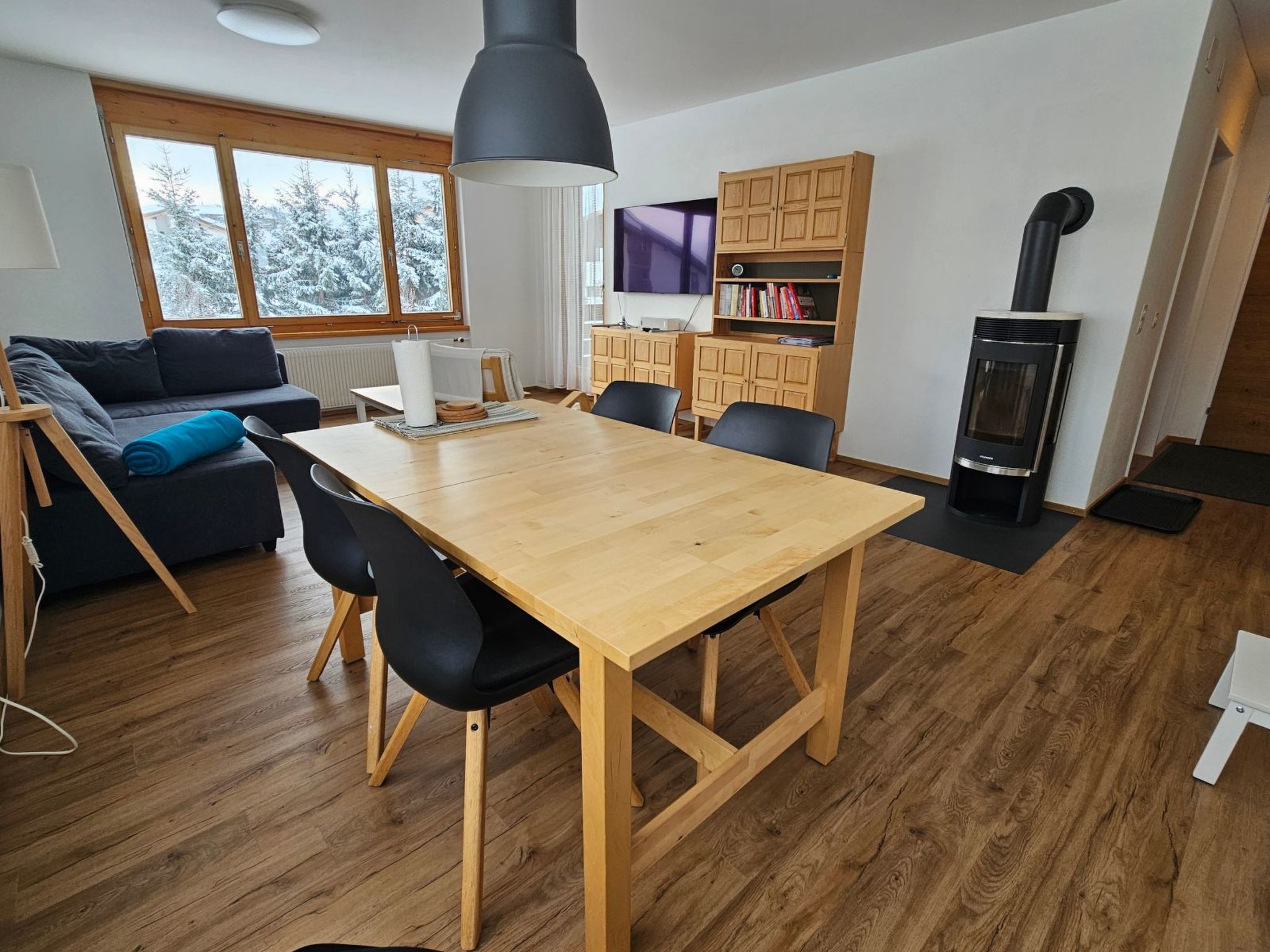 Ein moderner Wohnraum mit einem Holzesstisch, schwarzen Stühlen, einer Couch, einem Fernseher, Holzausbau und einem Kaminofen.
