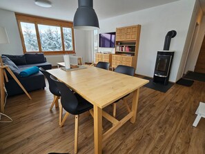 A modern living space with a wooden dining table, black chairs, a sofa, a television, wooden cabinets, and a wood-burning stove.