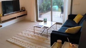 A modern living room with a TV, sofa, coffee table, and a view of a garden through a sliding door. - Sea & Elegance in the 8th district - Prado beaches on foot & garden terrace (Marseille)