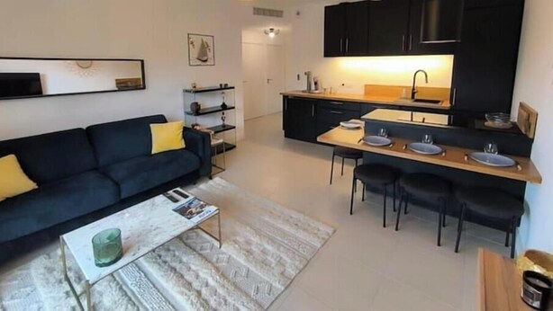 A modern living room with a sofa, coffee table, and a dining area with chairs and a kitchen with black cabinets and a sink. - Sea & Elegance in the 8th district - Prado beaches on foot & garden terrace (Marseille)