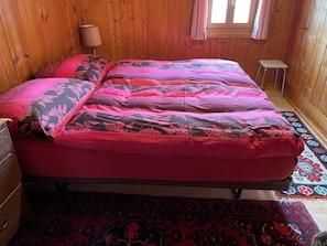 A bed with a patterned bedspread, a wooden floor, a window, and a small stool. - Venini, Rumein, (Degen), 45005B by Interhome (Vattiz)