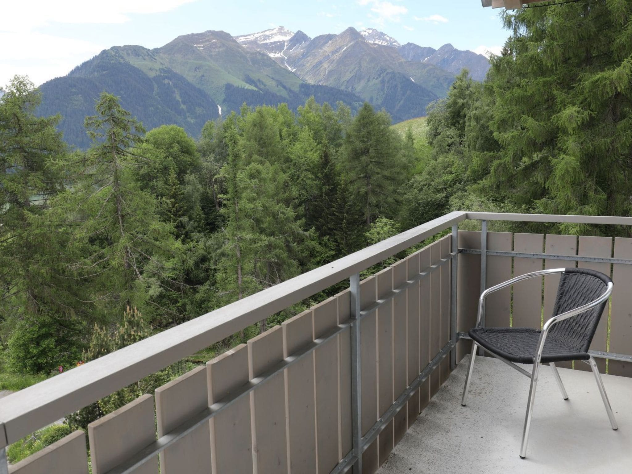 Ein Balkon mit Metallgeländer, einem einzelnen Stuhl und Blick auf eine gebirgige Landschaft mit dichtem Wald.
