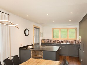 A modern kitchen with a central island, a dining area with a wooden table, and a view of a green landscape through large windows. - Vitg Vattiz, Vattiz, (Vattiz), 46002B by Interhome (Vattiz)