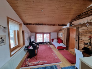 Ein gemütliches Zimmer mit Holzbalkendecke, einem Bett mit roter Bettwäsche, einem roten Teppich und einem kleinen Tisch mit einer Lampe.
