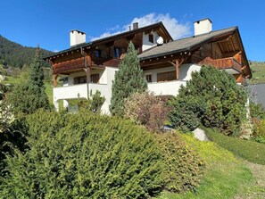 A two-story wooden chalet with a balcony, surrounded by dense greenery and a mountainous backdrop. - Casa Belinda Haht, Brigels, (Breil/Brigels), 65062B by Interhome (Breil/Brigels)