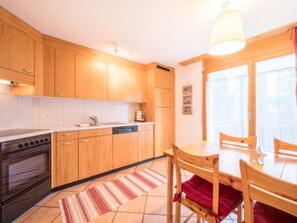 A kitchen with wooden cabinets, a stove, a dishwasher, and a dining area with a table and chairs. - Val Mulin 14.1 - Accornero by Interhome (Laax)