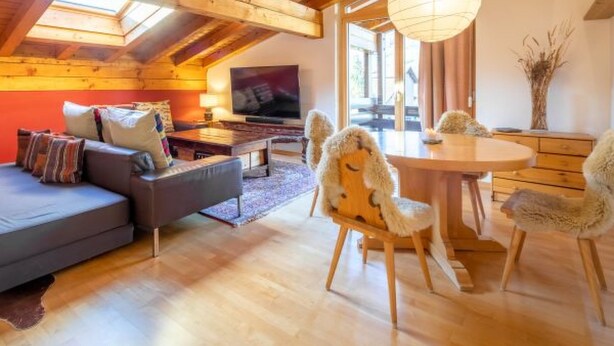 A cozy loft-style room with a wooden ceiling, a sofa, a TV, a dining table with chairs, and a large window. - Casa la Streila by Interhome (Laax)