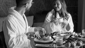 A couple in a robe seated at a table with breakfast items, including croissants and a cup of coffee, in a room with a textured wall.