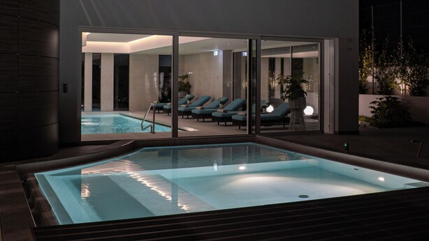 A modern indoor pool area with a glass-enclosed lounge area and a hot tub.